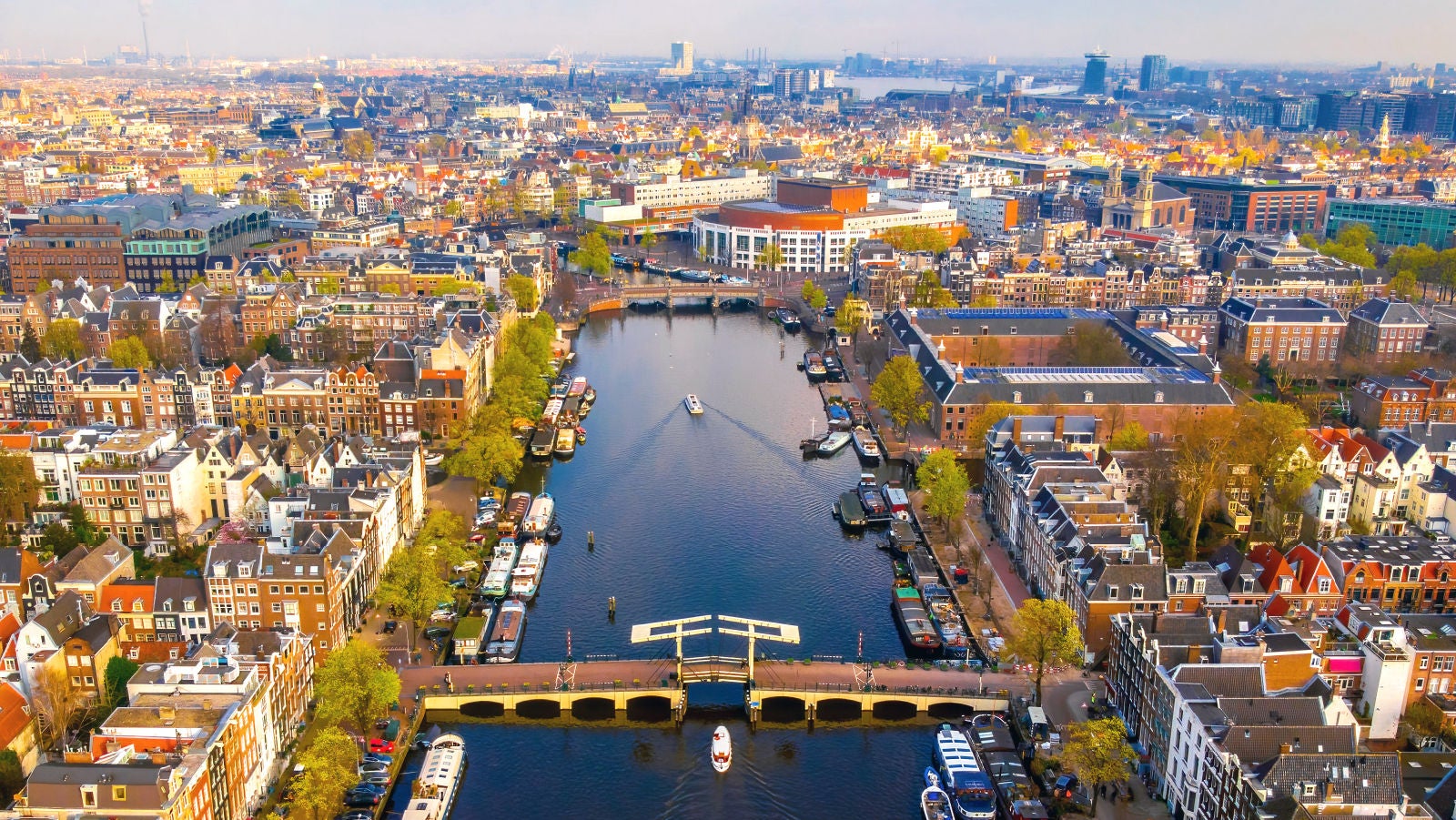Aerial view of Amsterdam, Netherlands with canals, bridges, boats, and dense historic buildings under a clear sky.