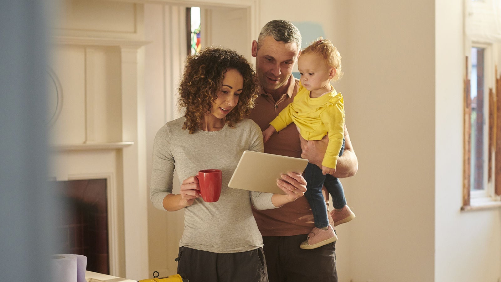 A couple standing in a home holding a tablet while the man carries a young child, all looking at the screen together.