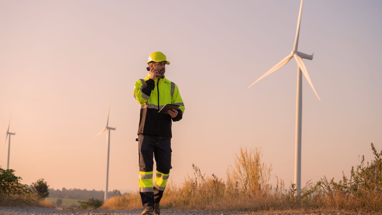 Utility worker in reflective safety clothing using a tablet and phone near wind turbines at sunset.