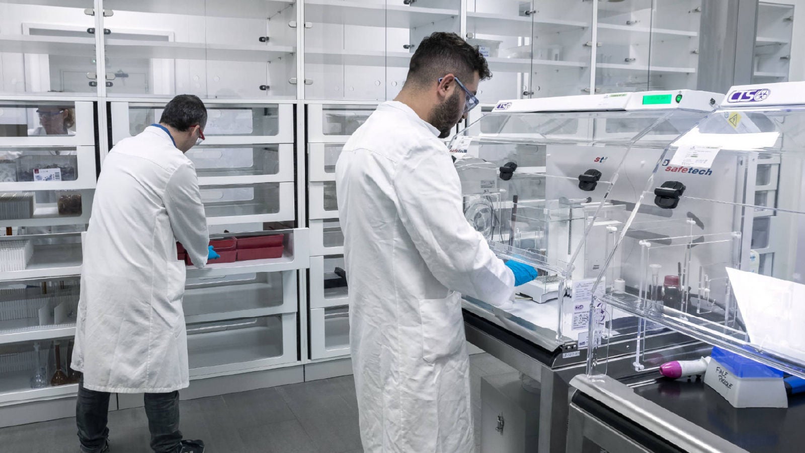Two laboratory technicians wearing white lab coats and safety glasses work with specialized equipment and storage units in a clean, modern lab environment.