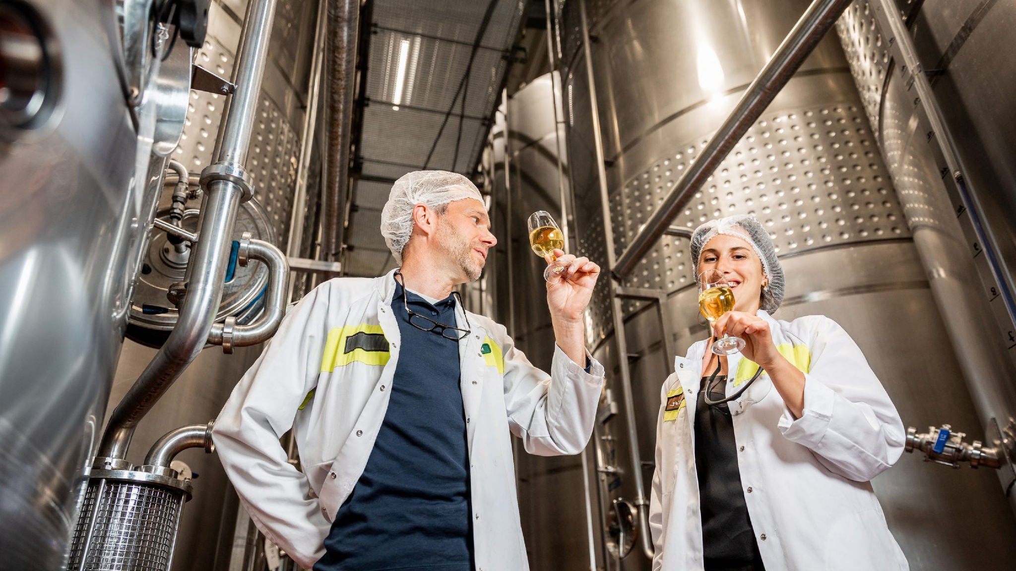 Two brewery employees wearing protective coats and hairnets inspecting glasses of wine inside a stainless steel production facility.