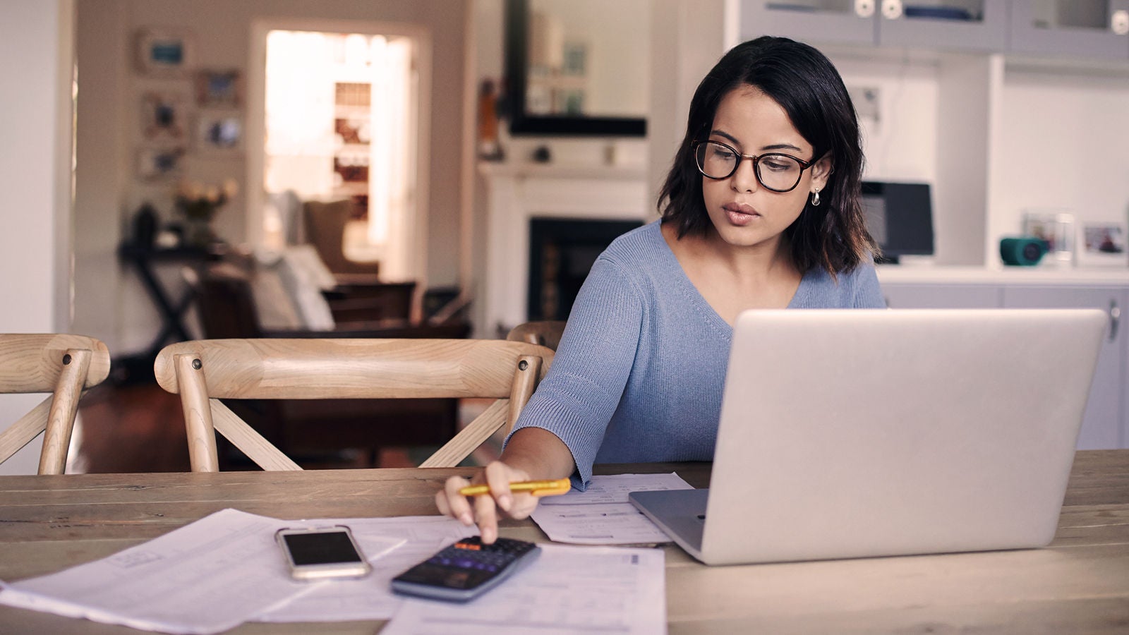The image shows a focused woman calculating and reviewing financial documents while working on a laptop at home, representing remote financial planning.