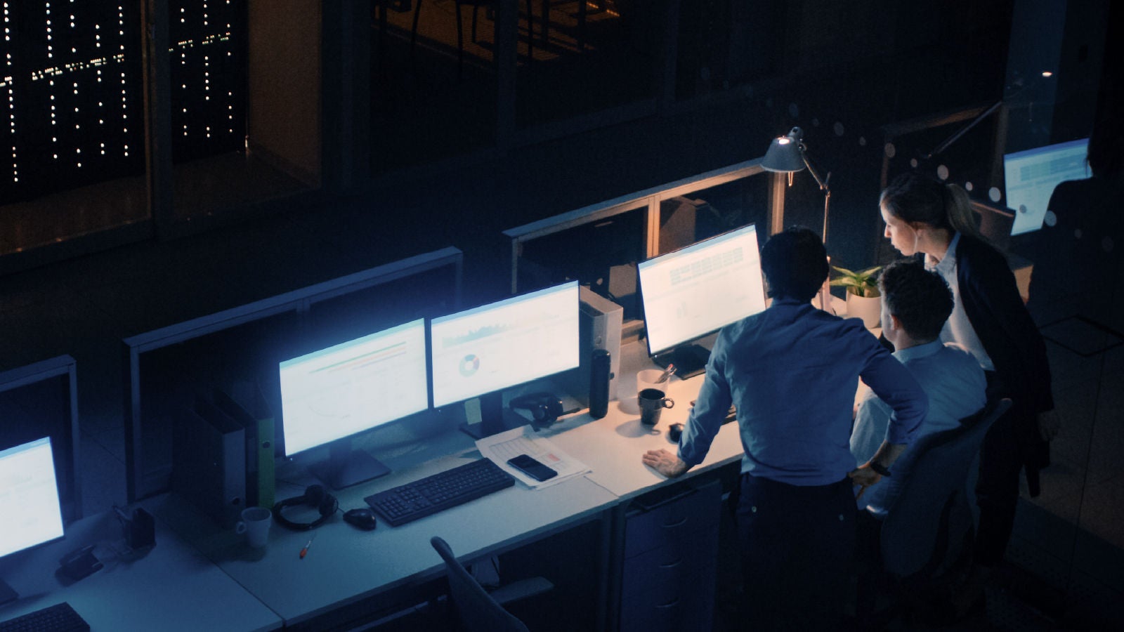 Three professionals in a dimly lit office working together at a desk with multiple glowing monitors.