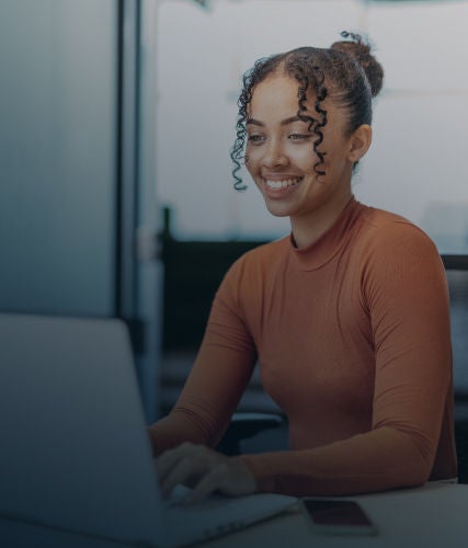 A smiling professional woman with her hair in a bun types on a laptop at a desk.