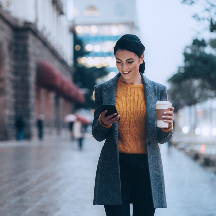 A woman in a yellow sweater and black pants holds a coffee while looking at her phone, appearing focused and relaxed.