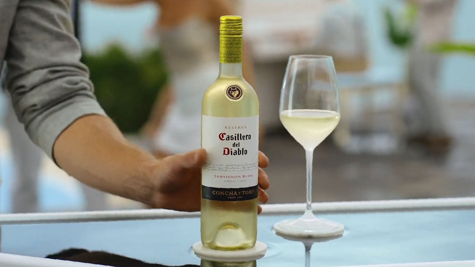 Man's arm grabbing a bottle of white wine next to a glass filled with white wine. 
