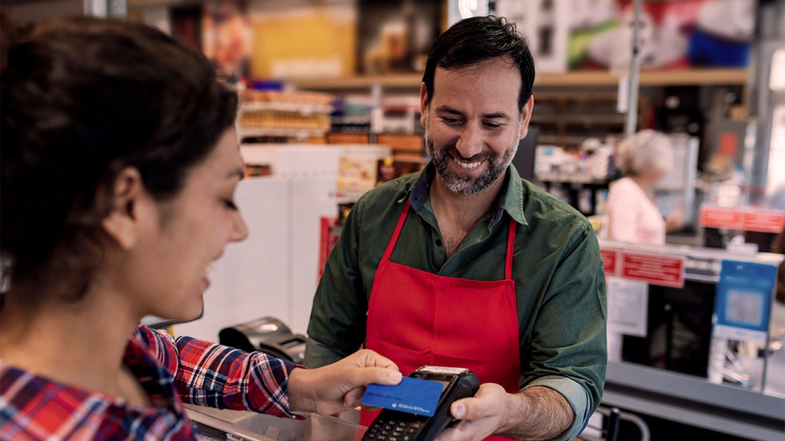 Smiling male cashier in a red apron holding a payment terminal while a female customer taps her card at a store counter.