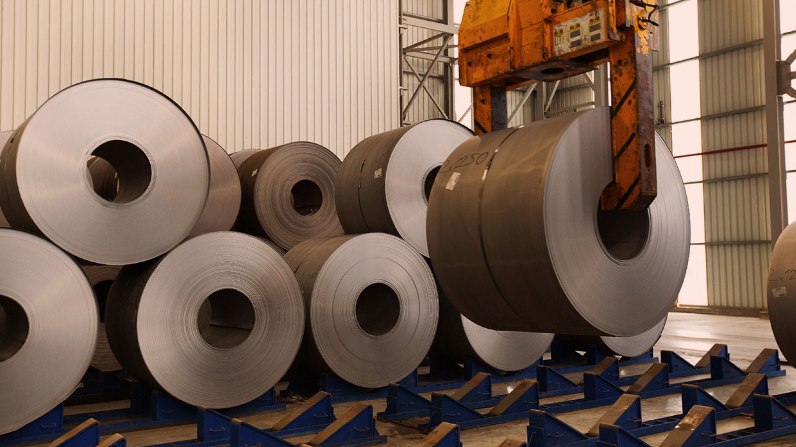 Large steel coils stacked inside an industrial warehouse with a crane lifting a metal coil.