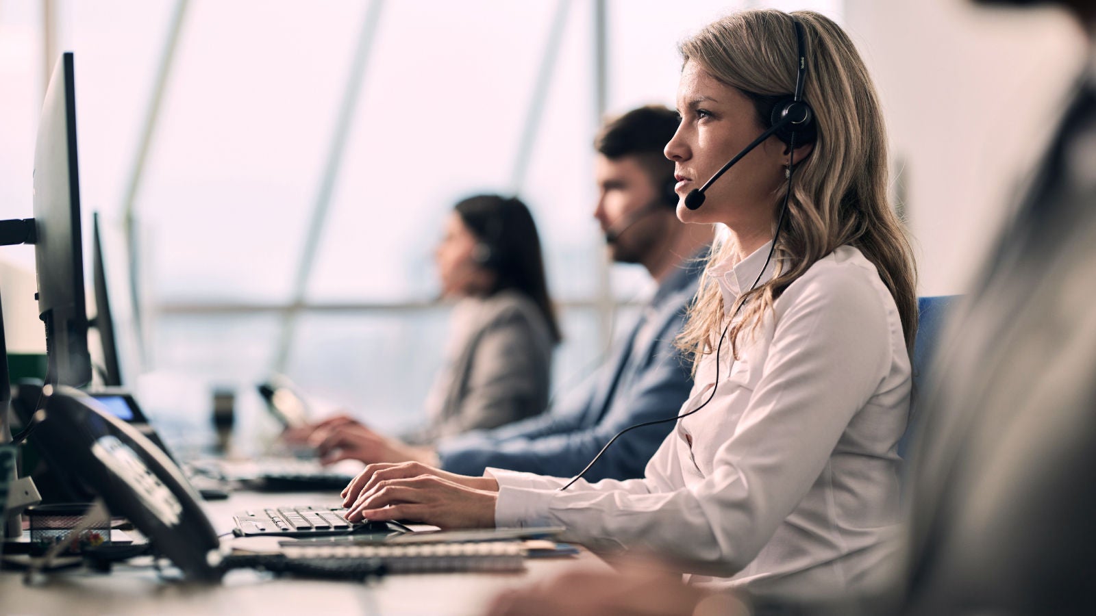 Customer support team seated in a row of desks, wearing headsets and working on computers in a bright office environment, representing collaboration and responsive service.