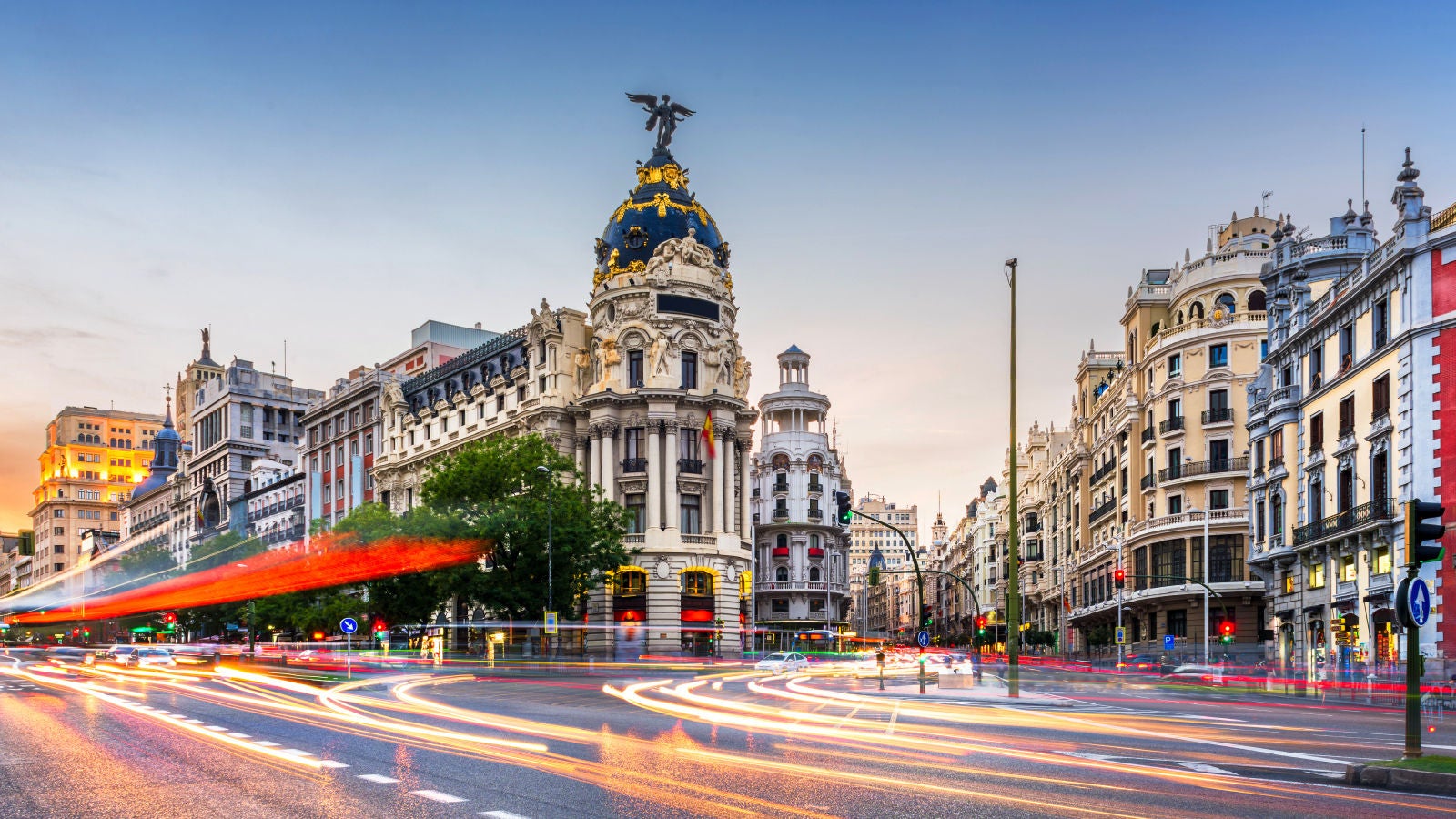Ornate historic buildings line a busy Madrid intersection with long-exposure traffic lights at dusk.