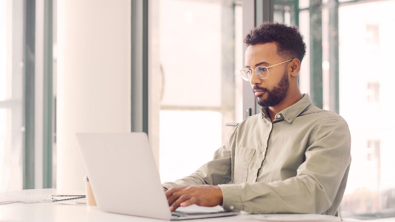 Professional in a light green button-up shirt sitting at a white desk, working on a laptop near large windows in a bright, modern office.