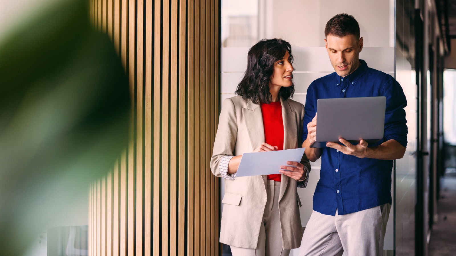 Two professionals standing in a modern office, engaged in discussion while looking at a laptop. One holds a document. Vertical wooden paneling and a glass wall frame the setting.