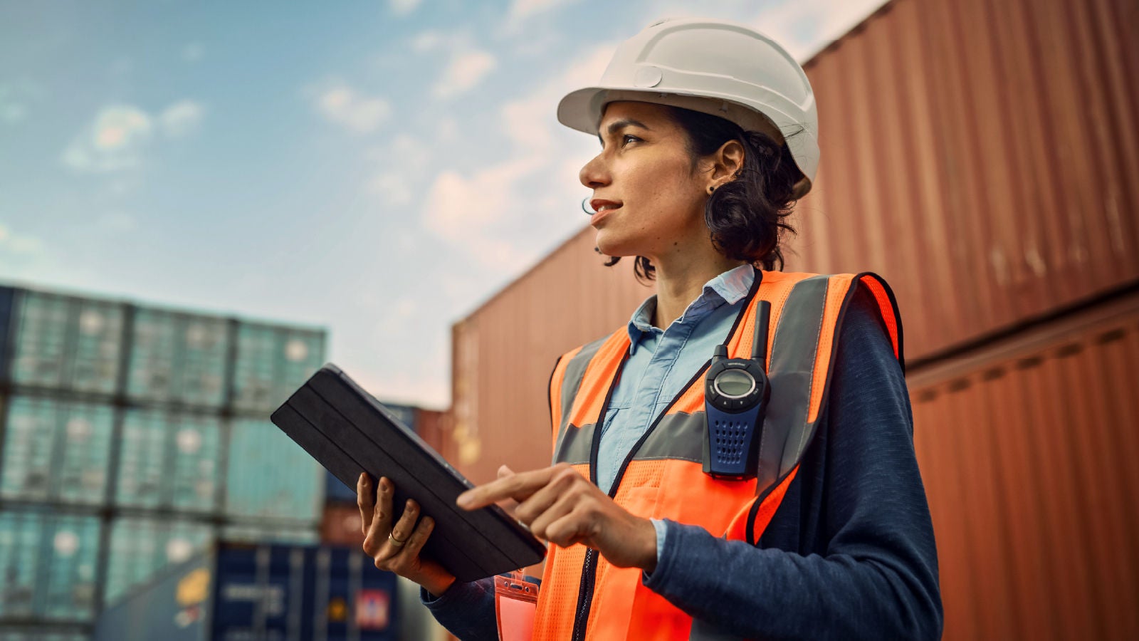 Female logistics worker wearing a hard hat and orange vest, holding a tablet in a shipping container yard under a clear sky.