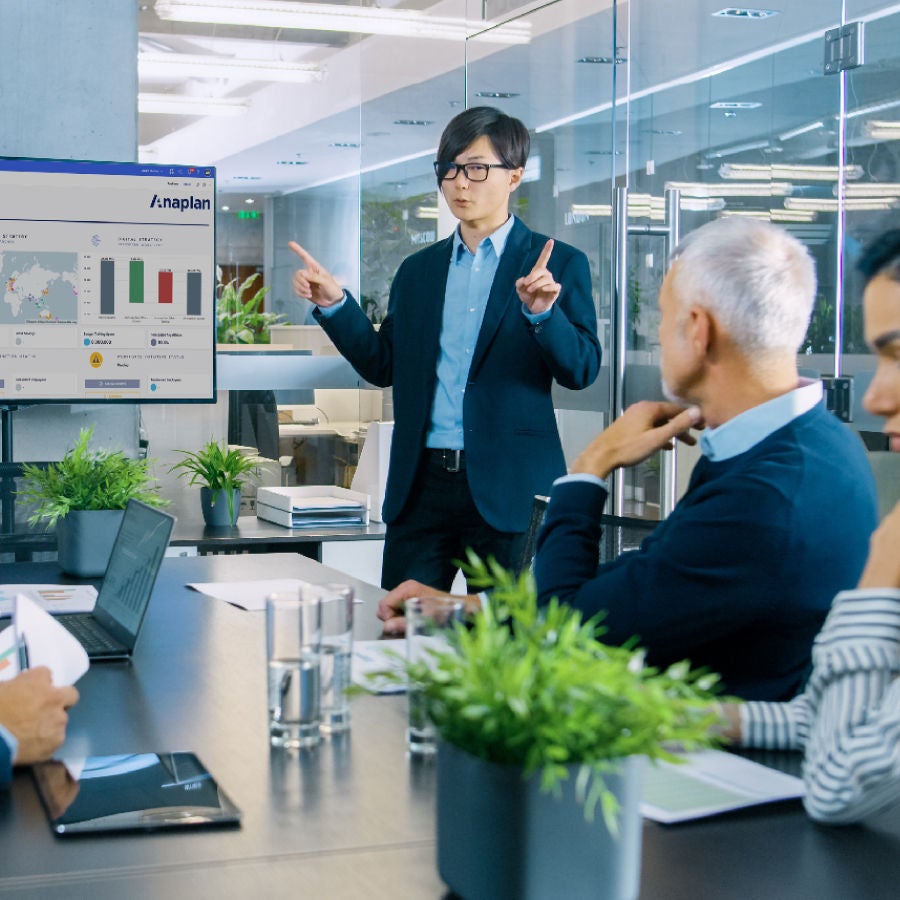 A man presents to a group of people in a meeting room, engaged in discussion and taking notes.