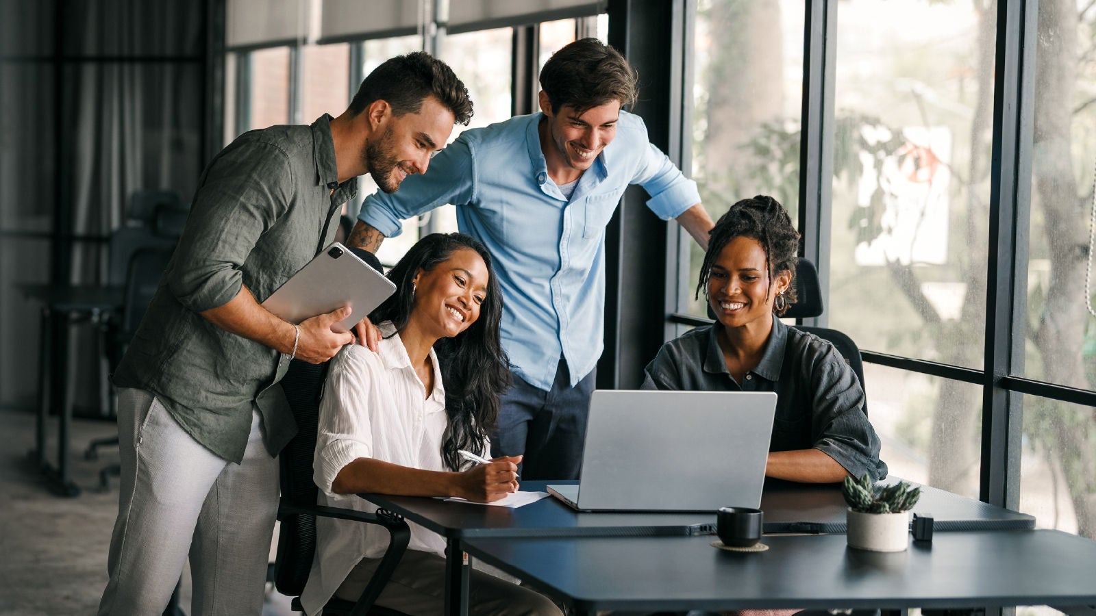 Four colleagues collaborate around a laptop in a bright modern office, smiling and reviewing work together.