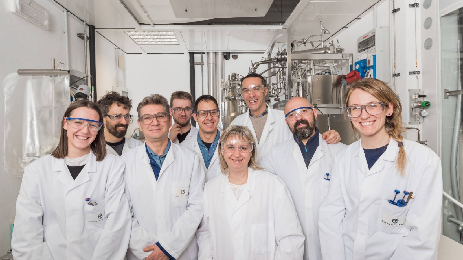 A group of scientists and researchers in white lab coats and safety glasses smiling together in a laboratory with stainless steel equipment and instruments in the background.