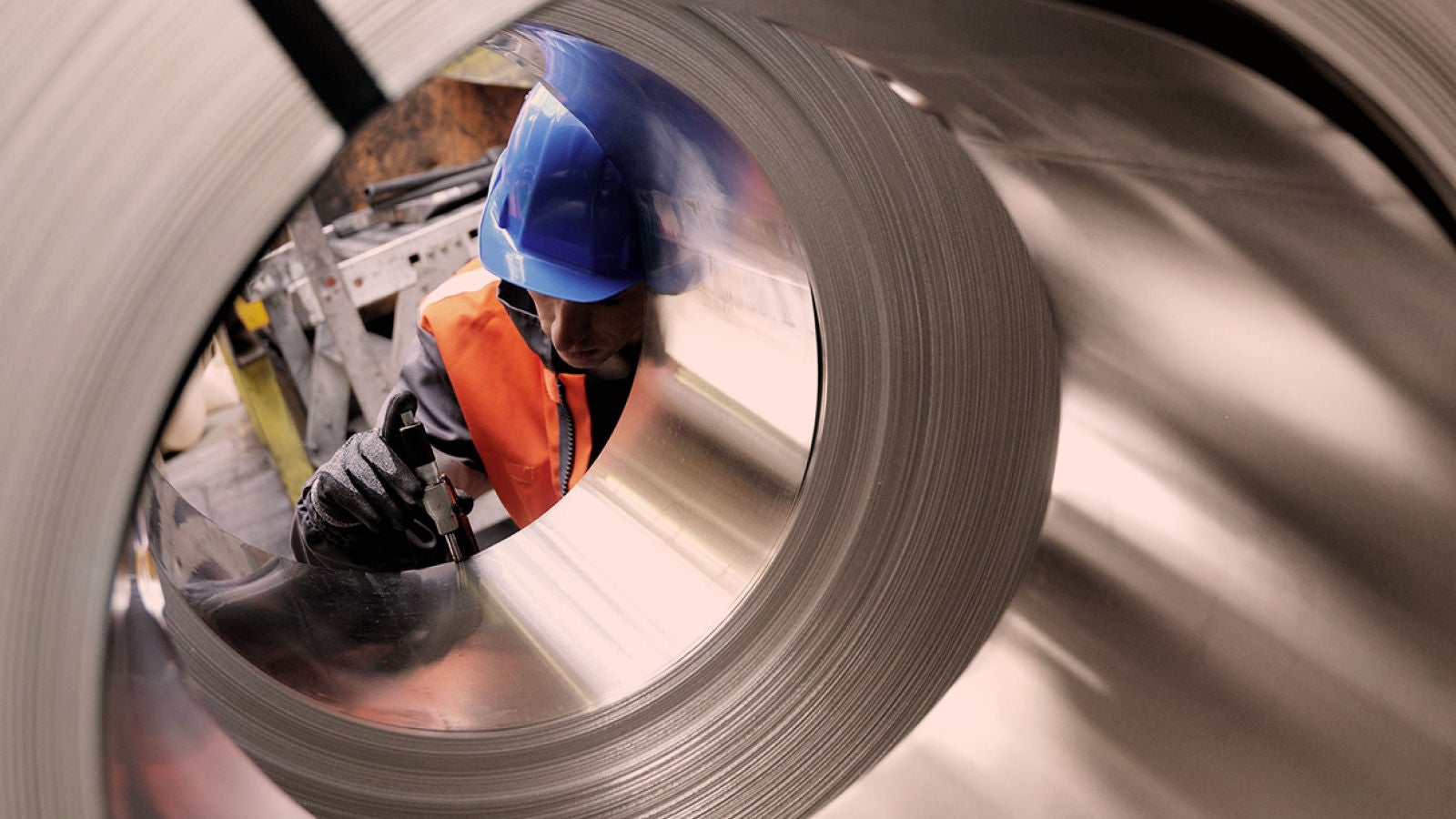 A worker wearing a blue hard hat and orange safety vest inspecting large rolled steel coils in a factory.
