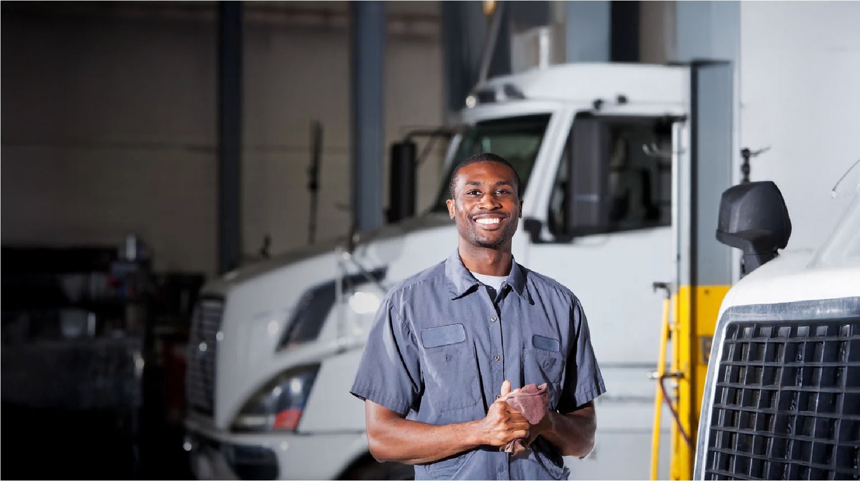 Smiling mechanic in a gray uniform holding a cloth, standing in a service garage with large white trucks in the background.