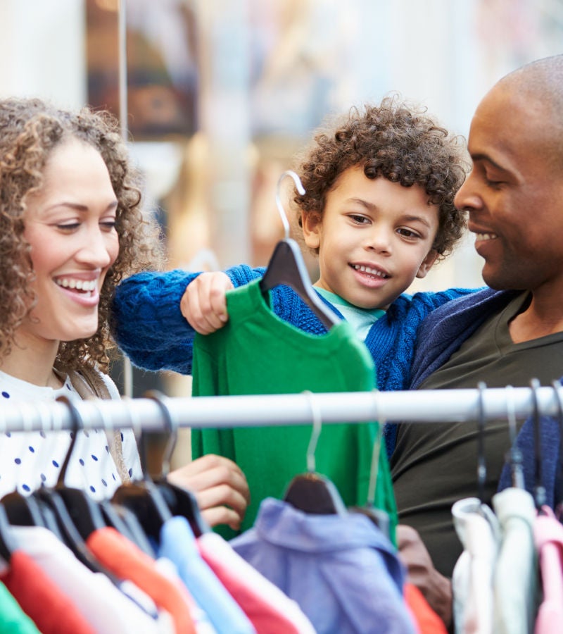 A smiling family shopping together, with a child holding up a green shirt on a hanger.