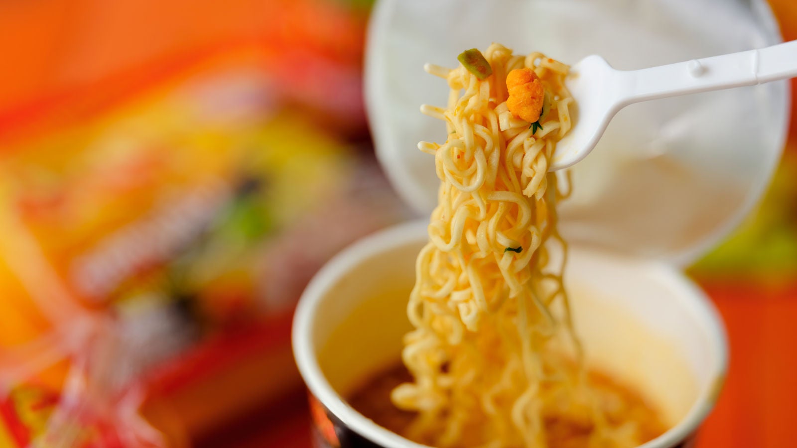 Instant noodles being lifted from a cup with a white plastic fork, with colorful packaging blurred in the background.