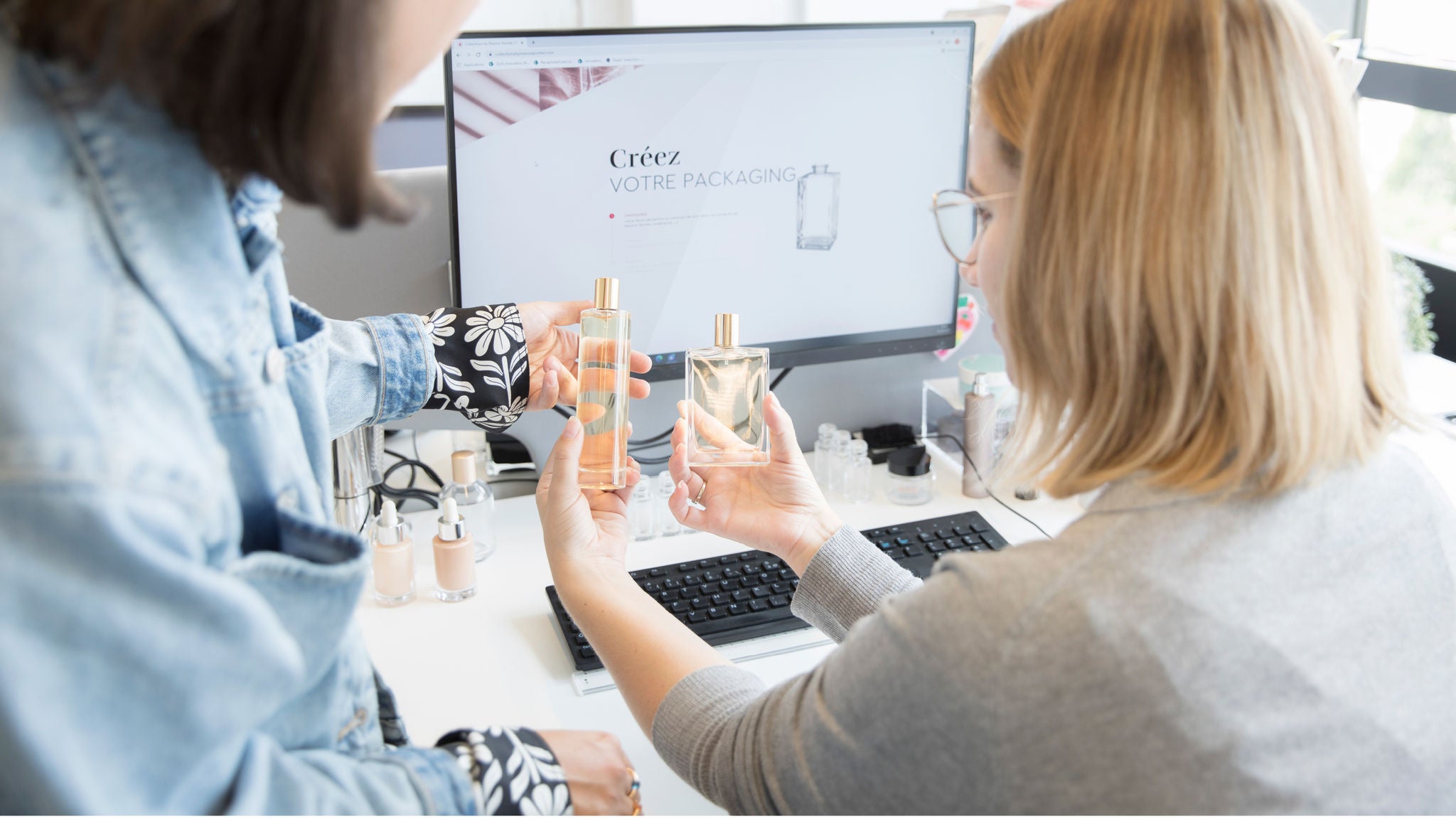 A woman in glasses shows perfume bottle samples to a colleague at a workstation.