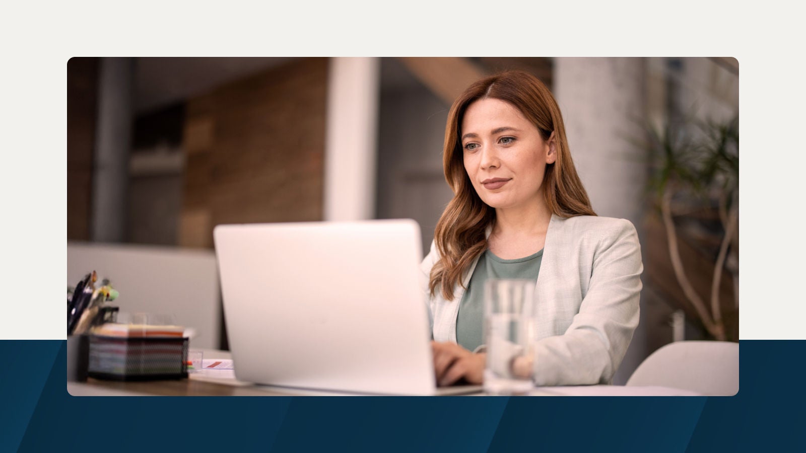 A professional businesswoman in a beige blazer working on a laptop in a modern office setting, with a blurred background.