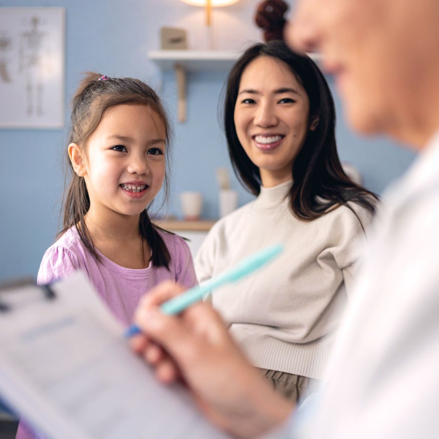 A young girl and her mother smile as a doctor writes notes on a clipboard during a medical appointment.