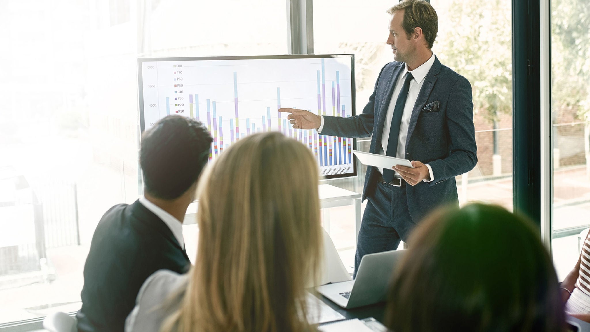 A business professional in a suit presents data on a large monitor displaying colorful bar graphs to a small group in a bright, modern office.
