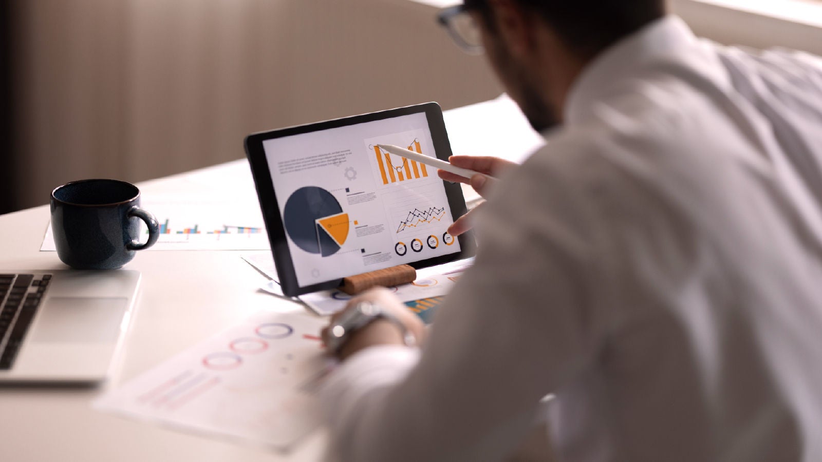 A man using a digital tablet to review financial pie charts, bar graphs, and trend lines at a desk with printed reports, a laptop, and a coffee mug.