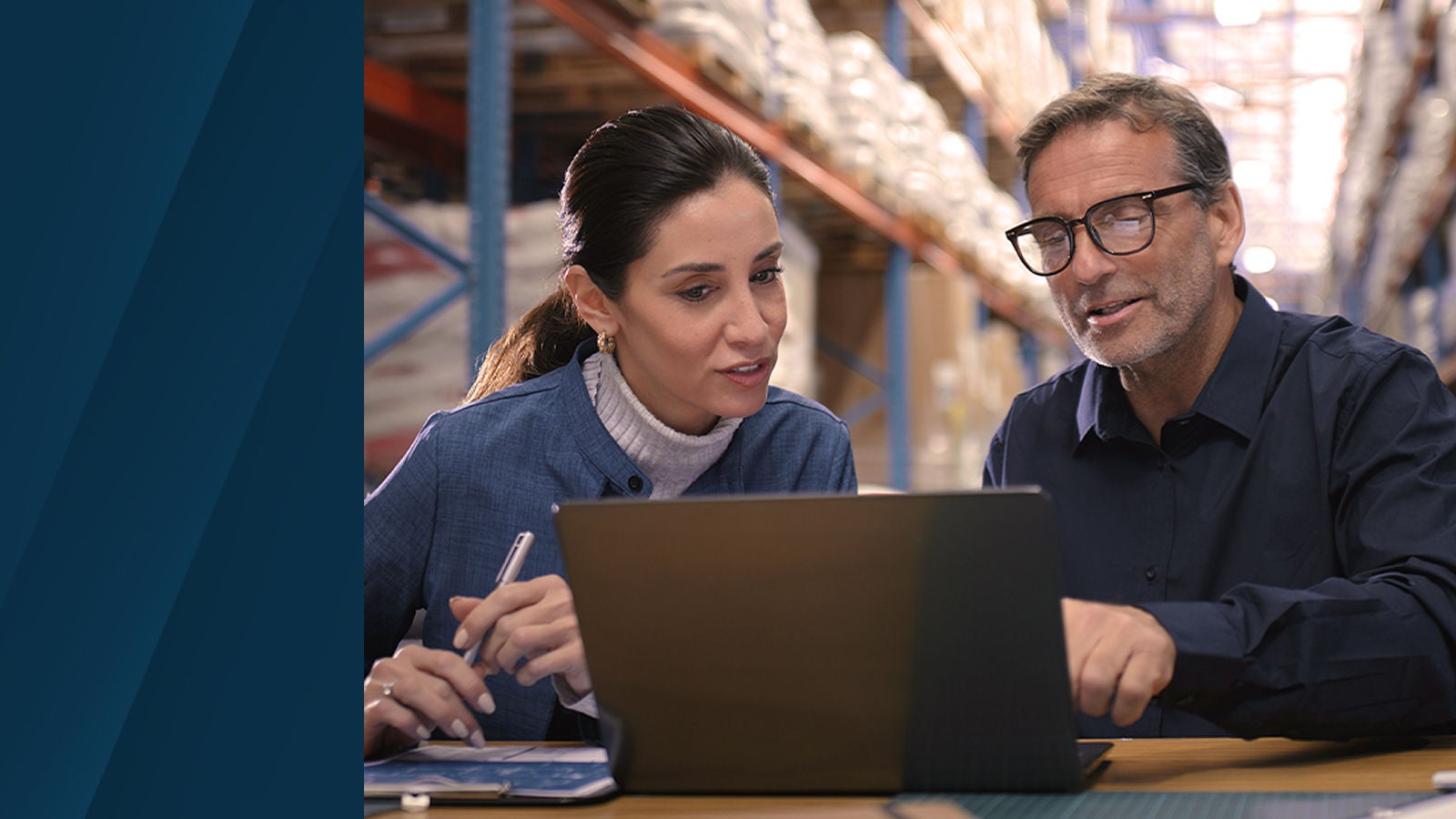 Two professionals seated at a table reviewing information on a laptop inside a warehouse environment, with shelving and inventory visible in the background.