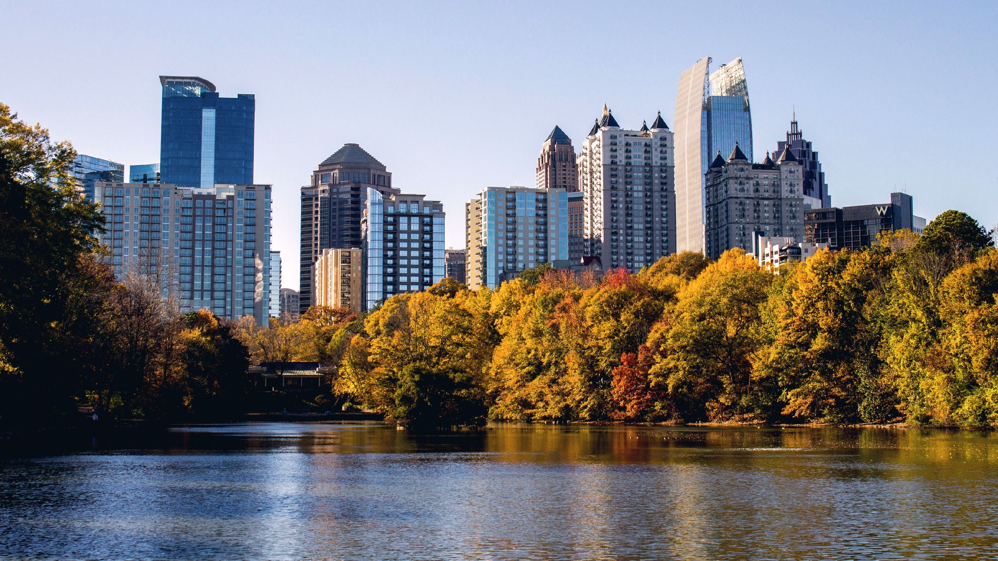 Atlanta city skyline with modern high-rise buildings behind a lake bordered by autumn-colored trees, reflecting on calm water under a clear blue sky.
