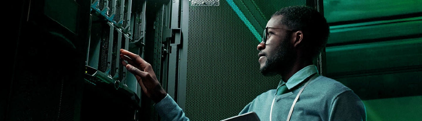 データ センターで技術者がノートパソコンを手にサーバー機器を調整している。緑のライトが点灯していて、ハイテクの雰囲気を醸し出している。