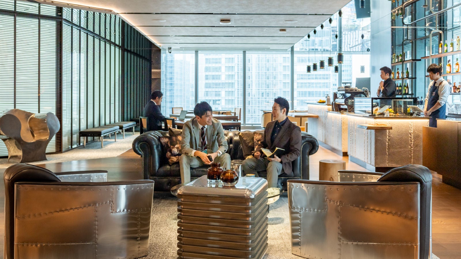Two business professionals converse while seated on a sofa in a modern office lounge with a café area in the background.