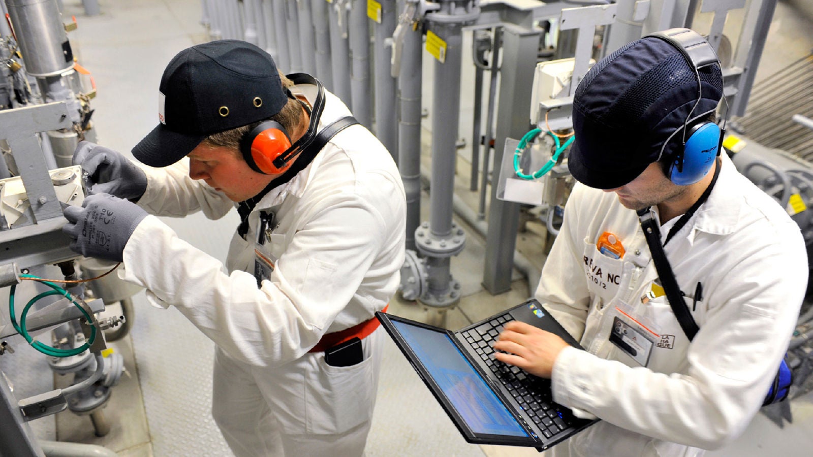 Two industrial technicians in protective clothing and ear protection work inside a control facility; one operates machinery while the other inputs data on a rugged laptop.