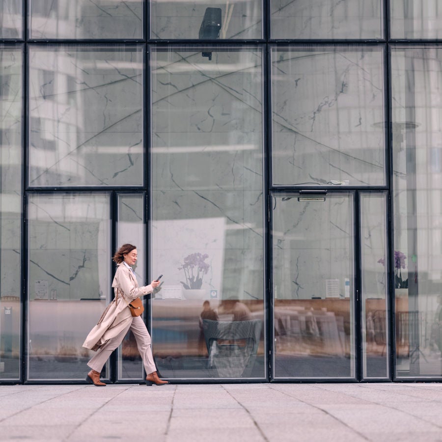A woman walks alongside a large glass building, showcasing modern architecture and city life.