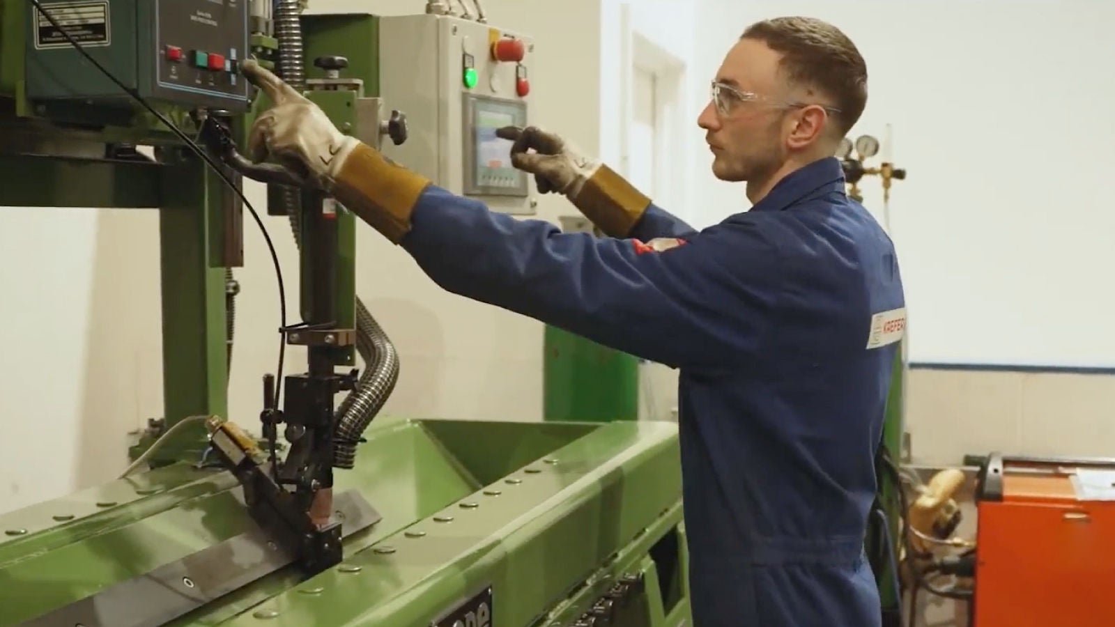 A technician in blue protective gear and gloves operates a control panel on green industrial machinery inside a clean, white facility.