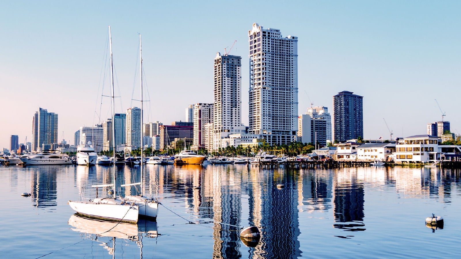 A marina in front of a modern city skyline with sailboats and yachts docked on calm water, reflecting tall buildings under a clear sky.