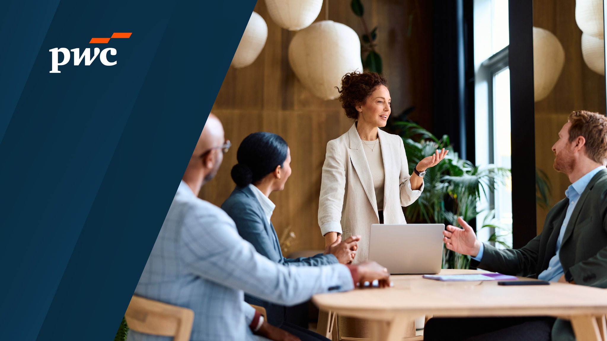 PwC logo on the left with a scene of a woman presenting to colleagues in a meeting room, standing beside a laptop while others sit at a table listening and discussing.