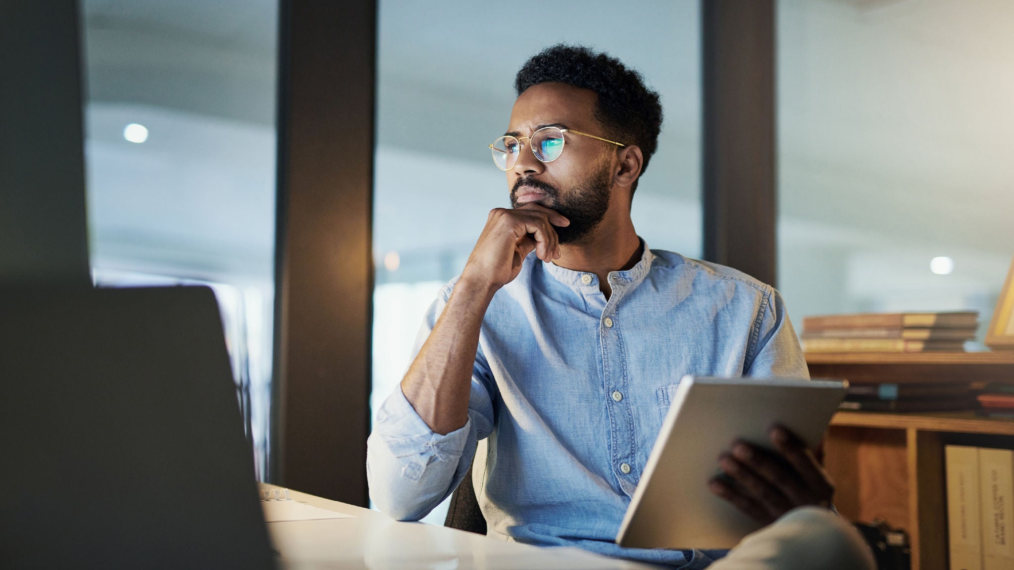 Man in glasses and a light blue shirt holding a tablet, seated at a desk with books in the background.