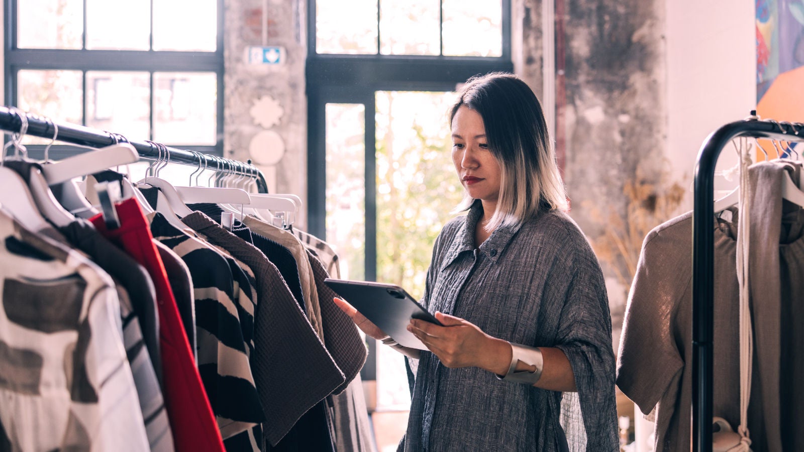 A woman in a retail store examines a tablet while standing beside a rack of clothing, with natural light streaming in through large industrial-style windows.