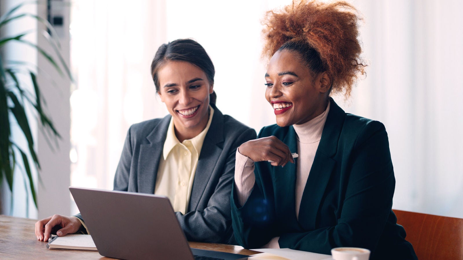 The image shows two women in business attire smiling and reviewing content on a laptop together in a bright office setting.