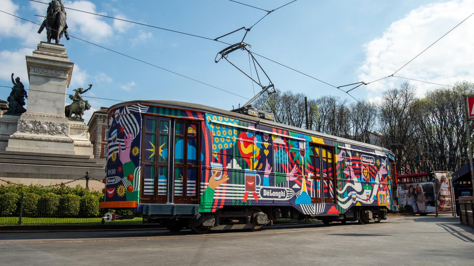 Colorful advertising-wrapped tram traveling past a city monument.