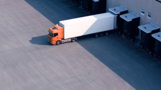 An orange truck parked in front of a large warehouse, showcasing its vibrant color against the industrial backdrop.