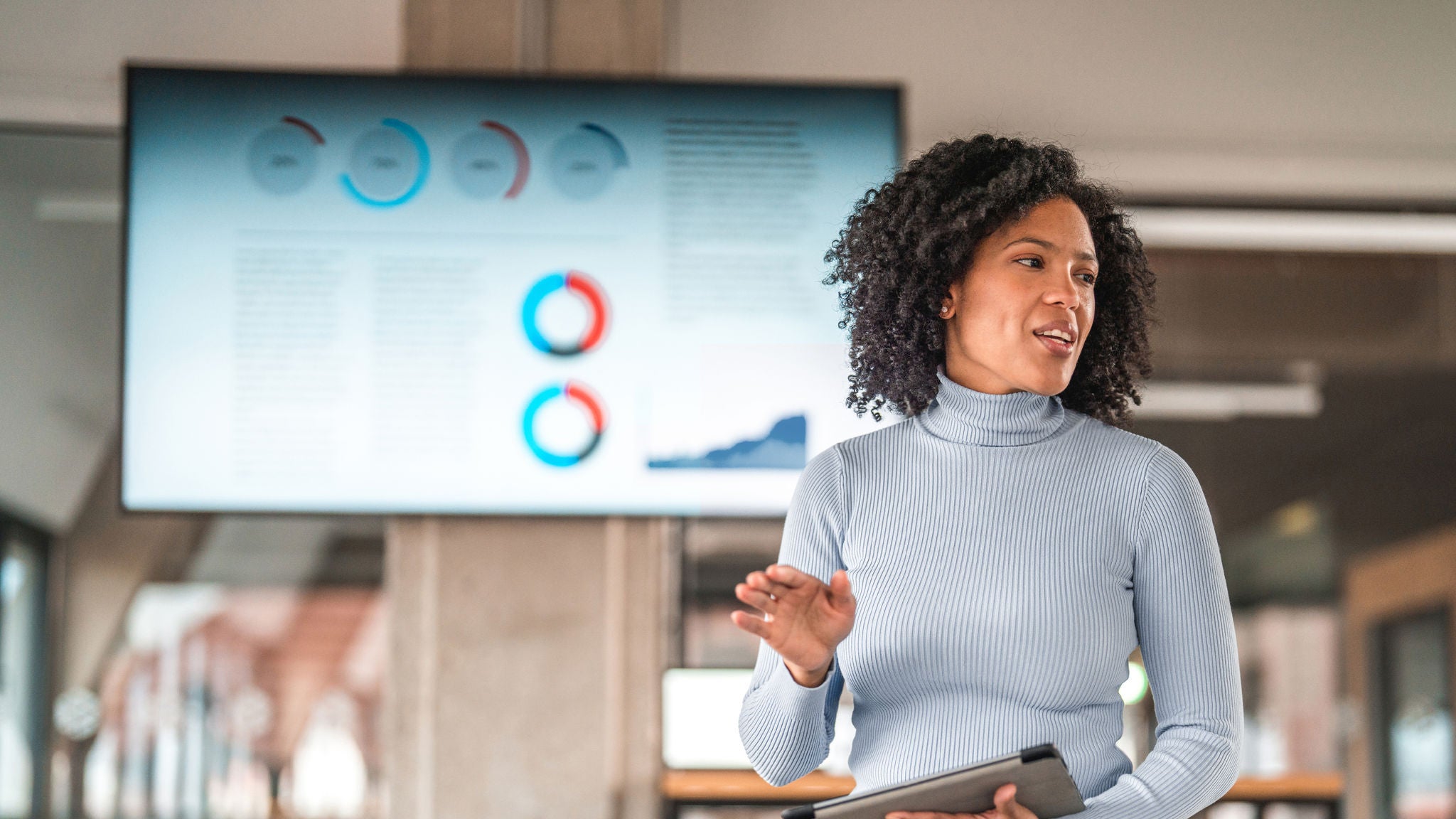 A woman holding a tablet while presenting in an office, with charts and graphs displayed on a screen behind her.