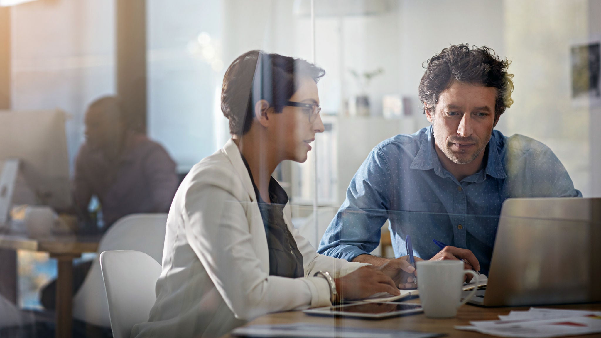 Two professionals reviewing data together on a laptop in a modern office environment, viewed through a glass wall.