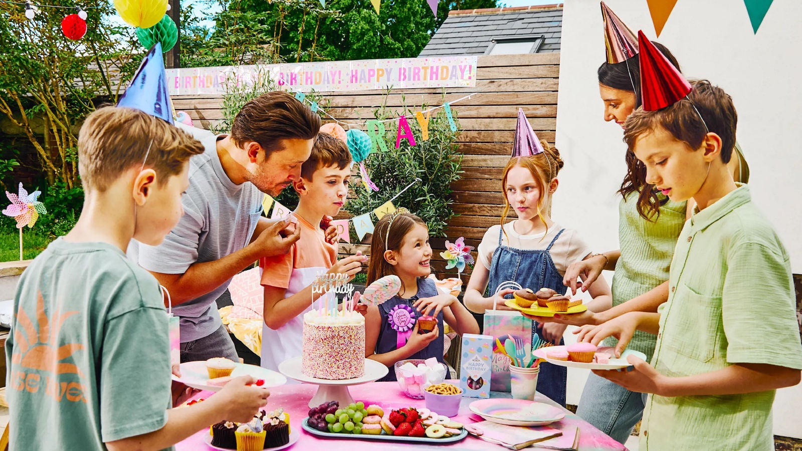 A cheerful outdoor birthday party with children and adults wearing party hats, enjoying cake and snacks around a decorated table.