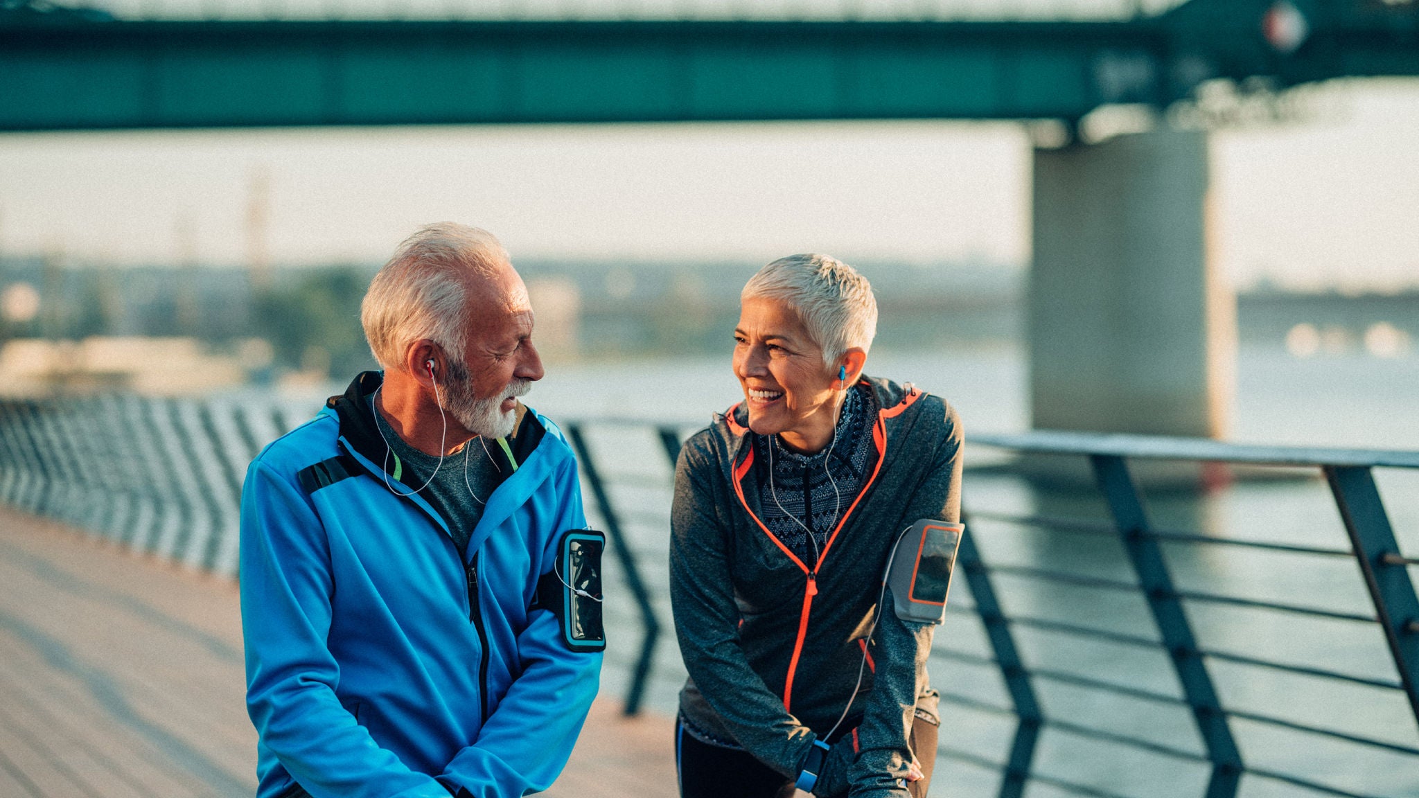 An older man and woman in athletic wear smiling and talking while cooling down after exercise near a waterfront walkway.