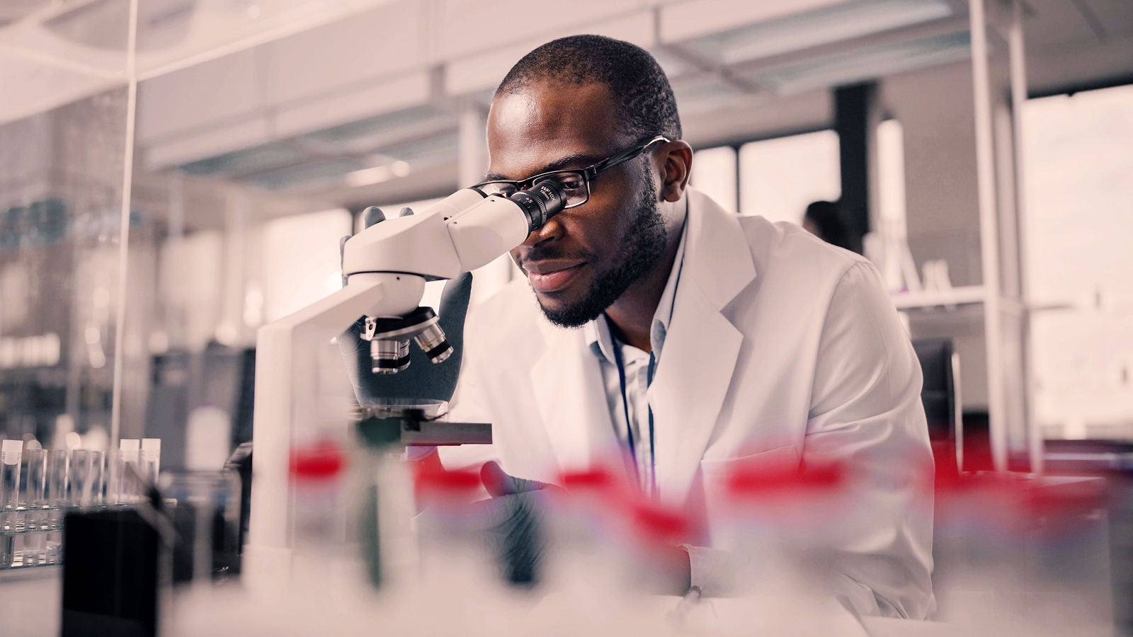 Scientist in a white lab coat looking through a microscope in a laboratory, with blurred test tubes in the foreground and shelves in the background.