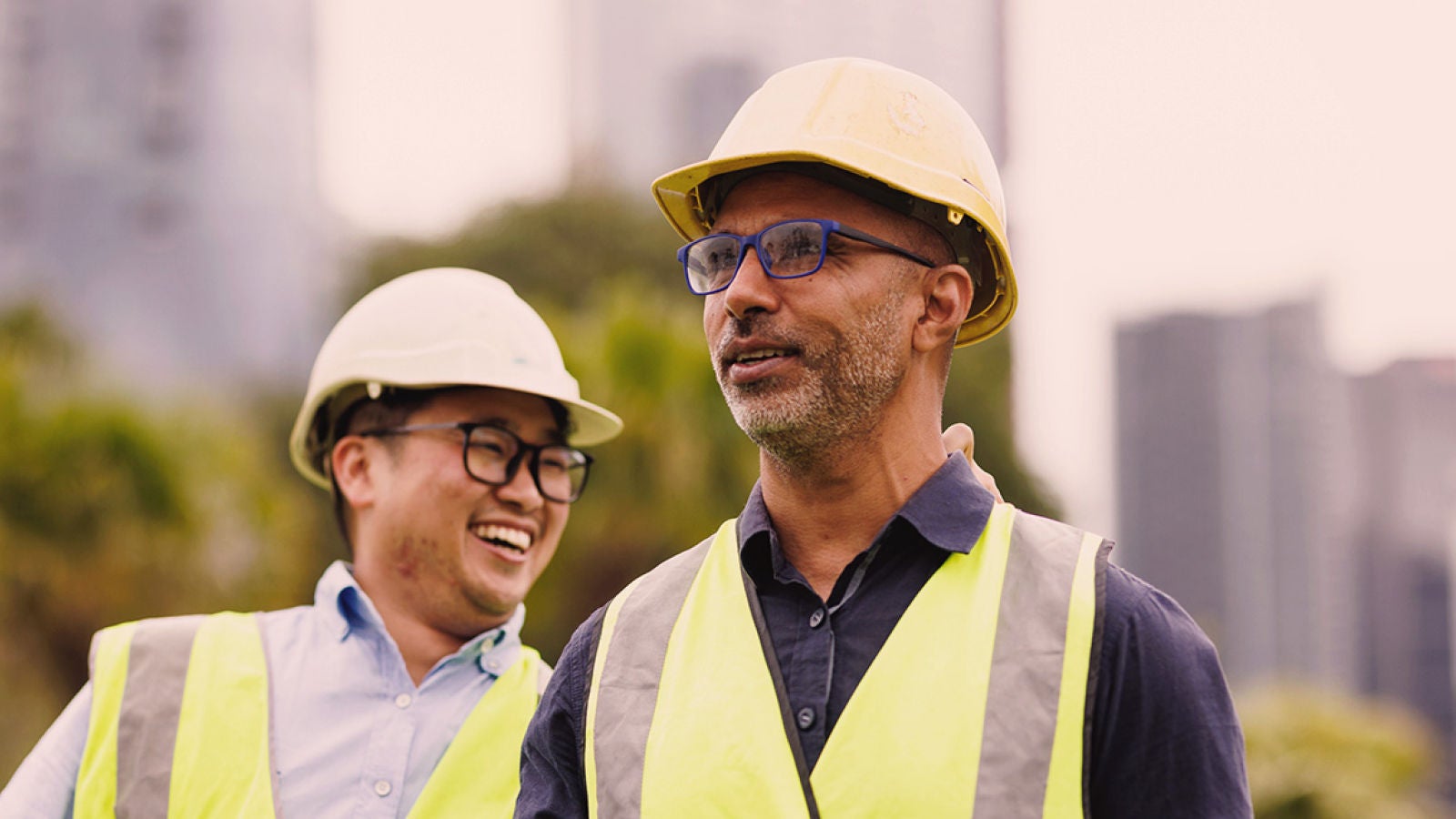 Two workers wearing hard hats and reflective safety vests standing outdoors at an industrial site.
