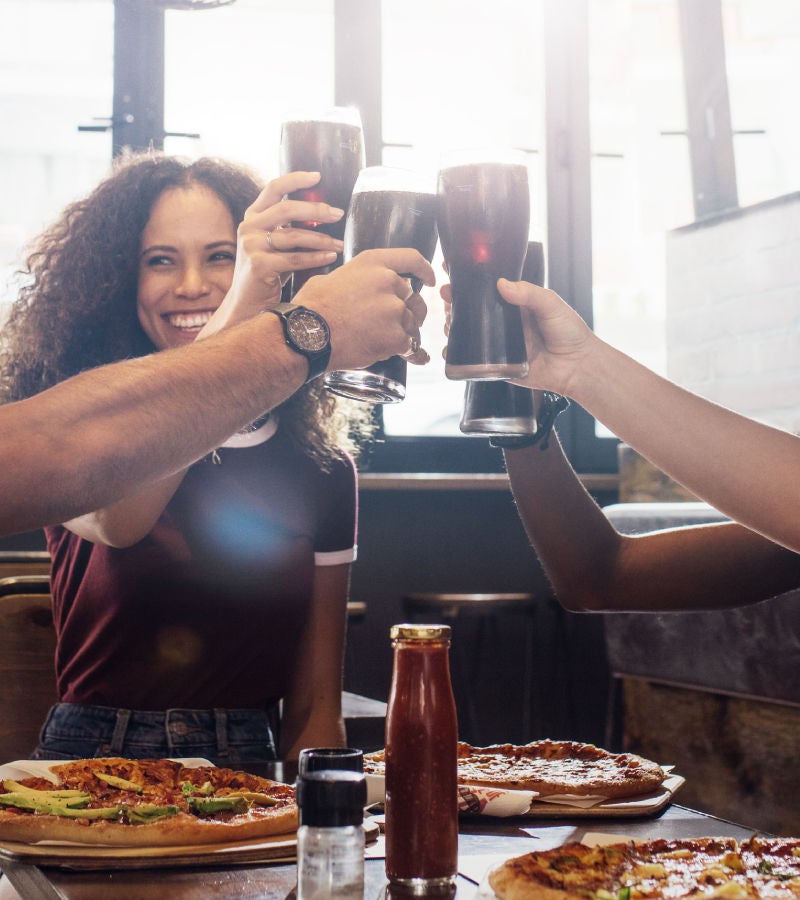 A group of friends raise glasses of soda in a toast, surrounded by pizzas and condiments at a restaurant table.